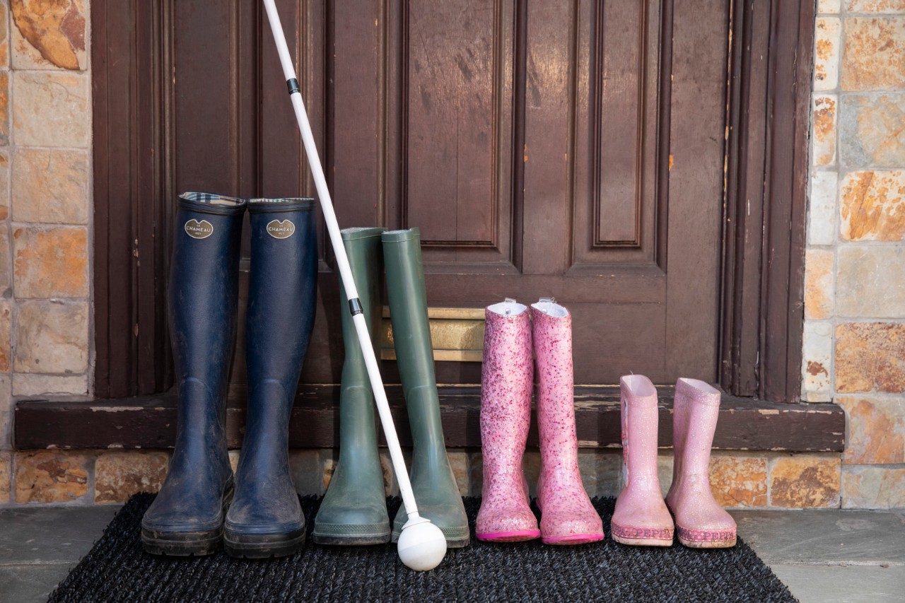four sets of colourful wellington boots sit lined up in descending order outside a front door with a long white ball cane alongside.