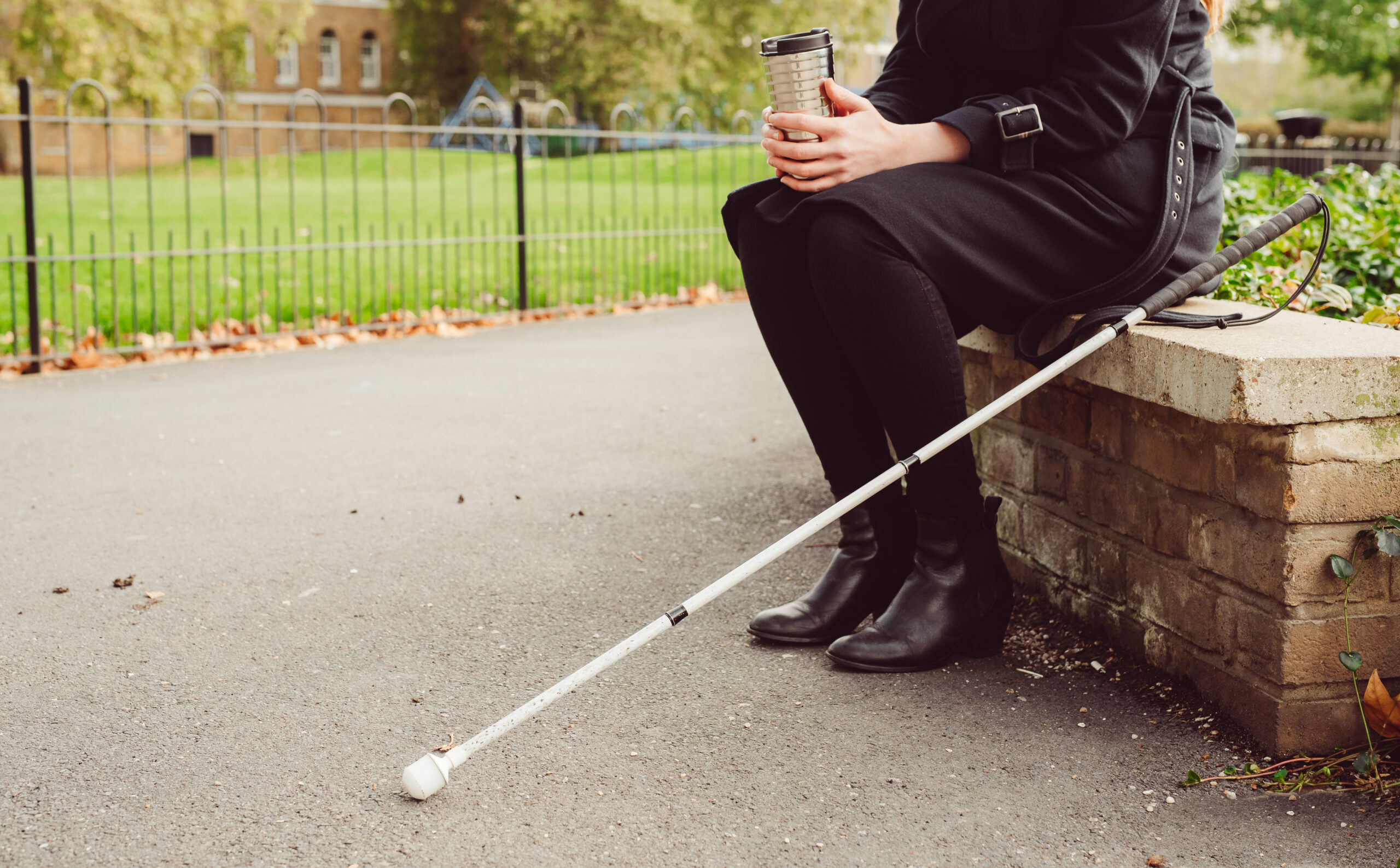 Bottom half of a woman sitting on a low wall, holding a coffee cup with a white cane beside her