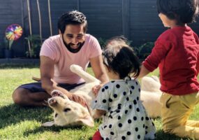 Image shows Amit playing with his two children and guide dog, Kika, in the garden.