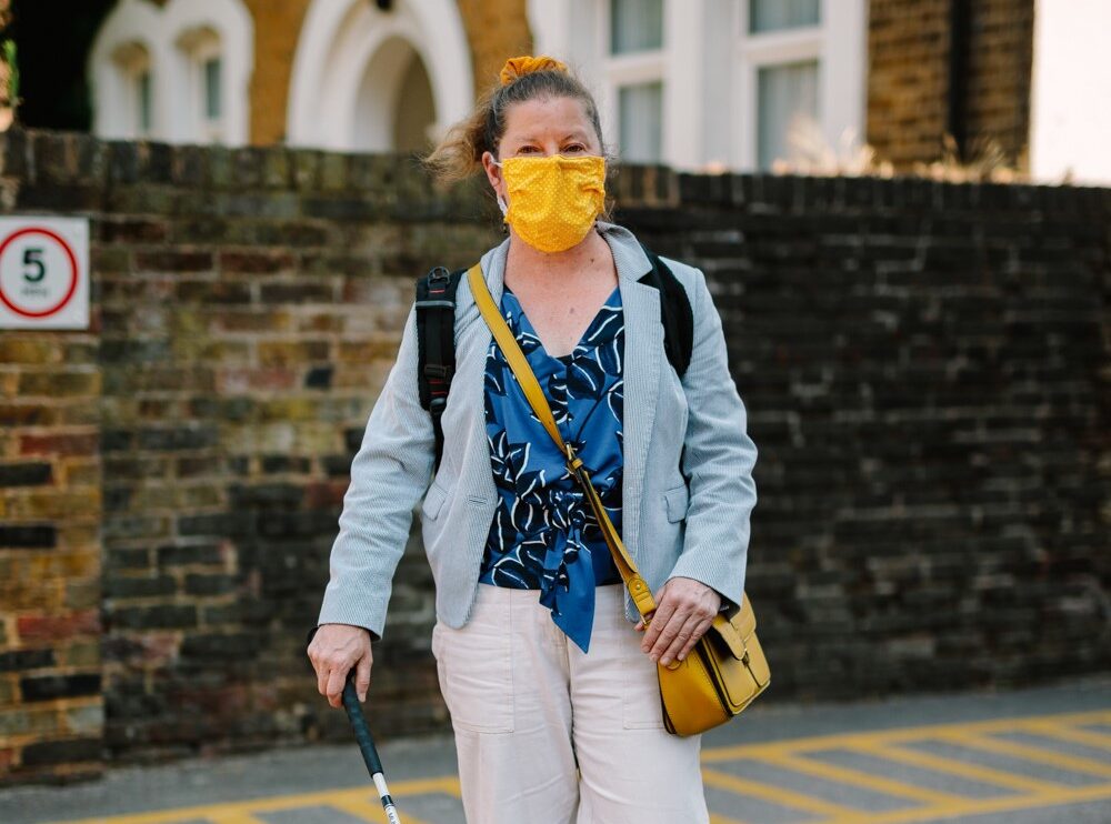A woman with a face mask walking down the street holding a white cane