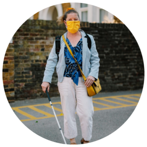 A lady, Odette, walking down the street with a white cane. Odette wears an orange face mask due to the Covid-19 pandemic.
