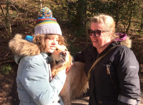 Image shows Chloe and her mum Jane smiling in the woods. Chloe is cuddling her golden buddy dog, Sapphire.