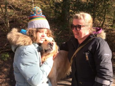 Image shows Chloe and her mum Jane smiling in the woods. Chloe is cuddling her golden buddy dog, Sapphire.