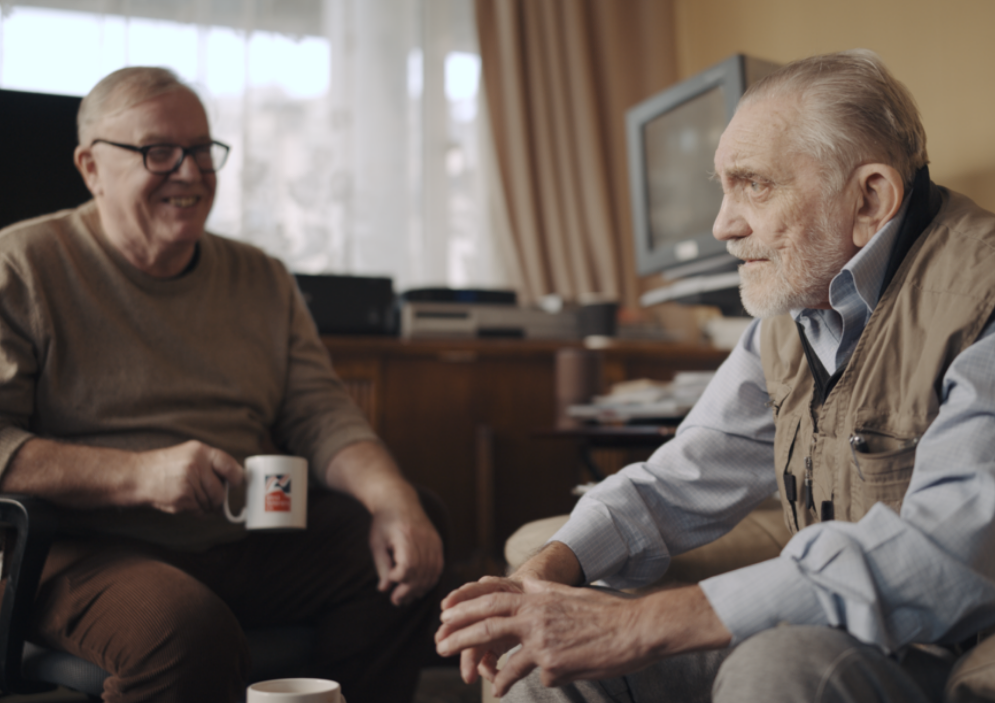 Image shows Ken talking to his support volunteer, Liam, in his living room. They smile over a cup of tea.