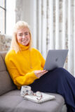 Becca a woman in her mid twenties with blond hair. She's wearing a bright yellow jumper and sitting cross legged on a sofa with a laptop balanced on her lap. Next to her is a mug, notebook and pair of glasses.