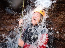Monica stands under a cascading waterfall, she's wearing a wetsuit and a yellow helmet. She is laughing and has her eyes closed.