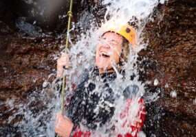 Monica stands under a cascading waterfall, she's wearing a wetsuit and a yellow helmet. She is laughing and has her eyes closed.