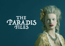 A production image of The Paradis Files: a young woman in 19th century costume stands in front of a team background. She has white hair piled atop her head and a very pale face with red lipstick