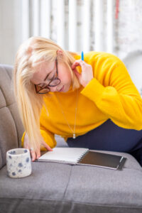Becca a blond woman wearing dark rimmed glasses is leaning over a notebook on a grey sofa. She's wearing a bright woollen jumper and has her hand behind her ear.