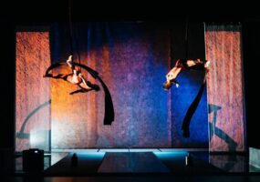 Flight Paths - A wide landscape shot of two women on aerial silks, tumbling through the air mid fall amongst a sea of orange and blue stage lighting.