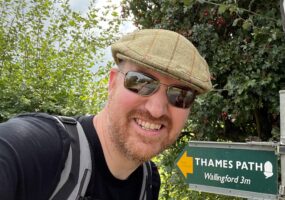Chris, a Caucasian man, stands smiling in front of a sign that says Thames Path. He is wearing sunglasses, a flat cap hat and a backpack.