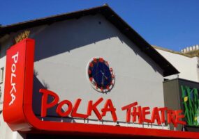 Outside the newly refurbished theatre. A white building with a triangular roof. Big red letters spell out Polka Theatre in fun script
