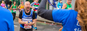 Team Vision runner, Duncan, cheering as he runs towards some Vision Foundation supporters in the crowd. There is a graphic of a calendar with “March 14th London Marathon ballot results day!” underlined.