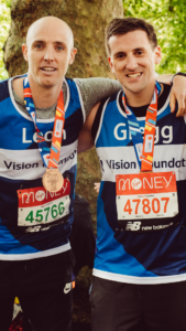 Leon and Gregg smiling arm in arm after the marathon. Both wearing Team Vision vests and their completion medals.