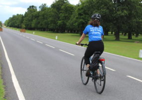 Cyclist on an open road in a park surrounded by leafy trees. The cyclist is photographed from behind and wearing a blue Vision Foundation t-shirt, reading “London’s sight loss charity”