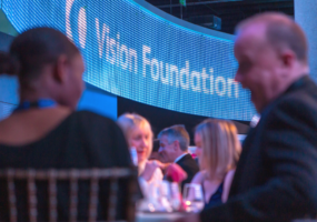 Two guests in conversation at their table. In the background there is an illuminated ellipse, featuring the Vision Foundation logo.