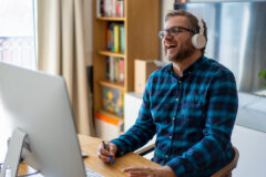 A white man with brown shaggy hair and a beard is laughing in front of a computer screen. He is wearing a blue checked shirt, white headphones and glasses.