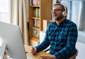 A white man with brown shaggy hair and a beard is laughing in front of a computer screen. He is wearing a blue checked shirt, white headphones and glasses.