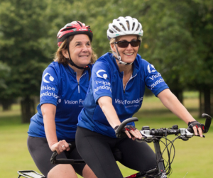 Monica and HRH The Countess of Wessex smiling widely whilst cycling on a tandem together