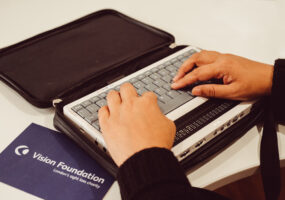 A woman's hands rest on a Braille keyboard. Next to her keyboard is a postcard with the words Vision Foundation on it.