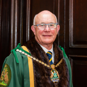 Nigel, a middle-aged white man. He is wearing a green and yellow cloak and a medallion around his neck as a Master Spectacle Maker