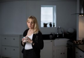 A blond woman stands in a kitchen holding a mug of tea and looks down at the floor