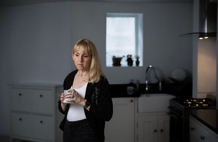 A blond woman stands in a kitchen holding a mug of tea and looks down at the floor