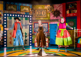 Three actors on stage, they wear colourful costumes. One wears a pirate inspired outfit, one in brown velvet dungarees and a princess in a tiered neon dress. They stand against a mix of colourful designs as a backdrop.