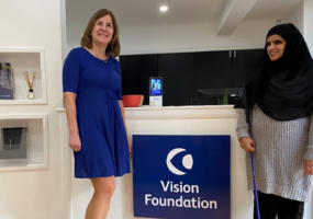 Monica and Khafsa stood beside Vision Foundation's Head Office reception. There is a Vision Foundation branded sign on the front of the reception desk.
