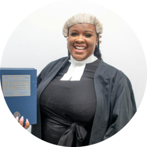 Jessikah Inaba, a black and blind woman in her barrister robe and wig. She is smiling and holding up a copy of the criminal practice book.