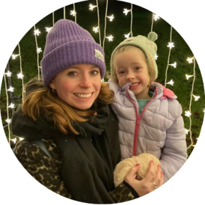 Fran and daughter, Maisie wrapped up in coats and woolly hats as they smile in front of a curtain of fairy lights.