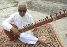 Baluji playing an instrument. seated cross legged on a carpet.