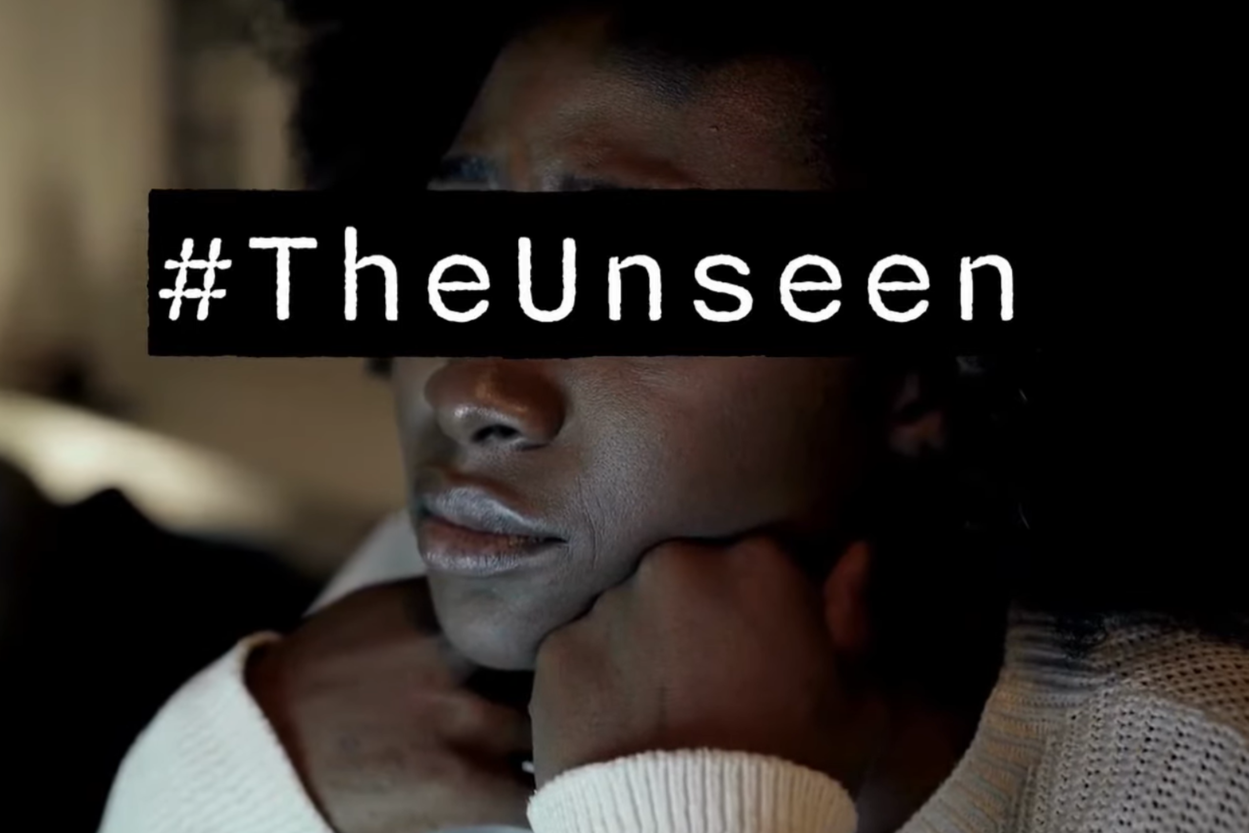 Image from The Unseen video featuring a lady resting her chin on her hands looking deflated. The room behind her is dark. #TheUnseen text in white on a black rectangle covers her eyes to give the lady anonymity.