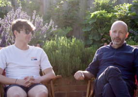 Seb, an 18year old white man with brown hair, sunglasses, a white shirt and shorts sits next to his father, Alan wearing a navy blue shirt and blue trousers. Who is in his 50s. They are sat in the sun behind trees and purple flowers.