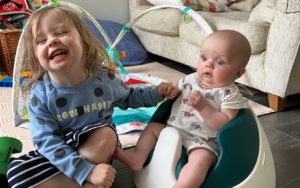 Harper, a blonde 3-year-old girl grinning for the camera. She is sat beside her baby brother, Jesse who is in a baby-set and they are holding hands.