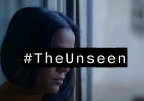 A still from our video about The Unseen Report. It features a lady with brown skin and dark hair looking out of a window. Her eyes are covered by the words #TheUnseen to make her anonymous.