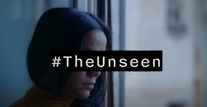 A still from our video about The Unseen Report. It features a lady with brown skin and dark hair looking out of a window. Her eyes are covered by the words #TheUnseen to make her anonymous. 