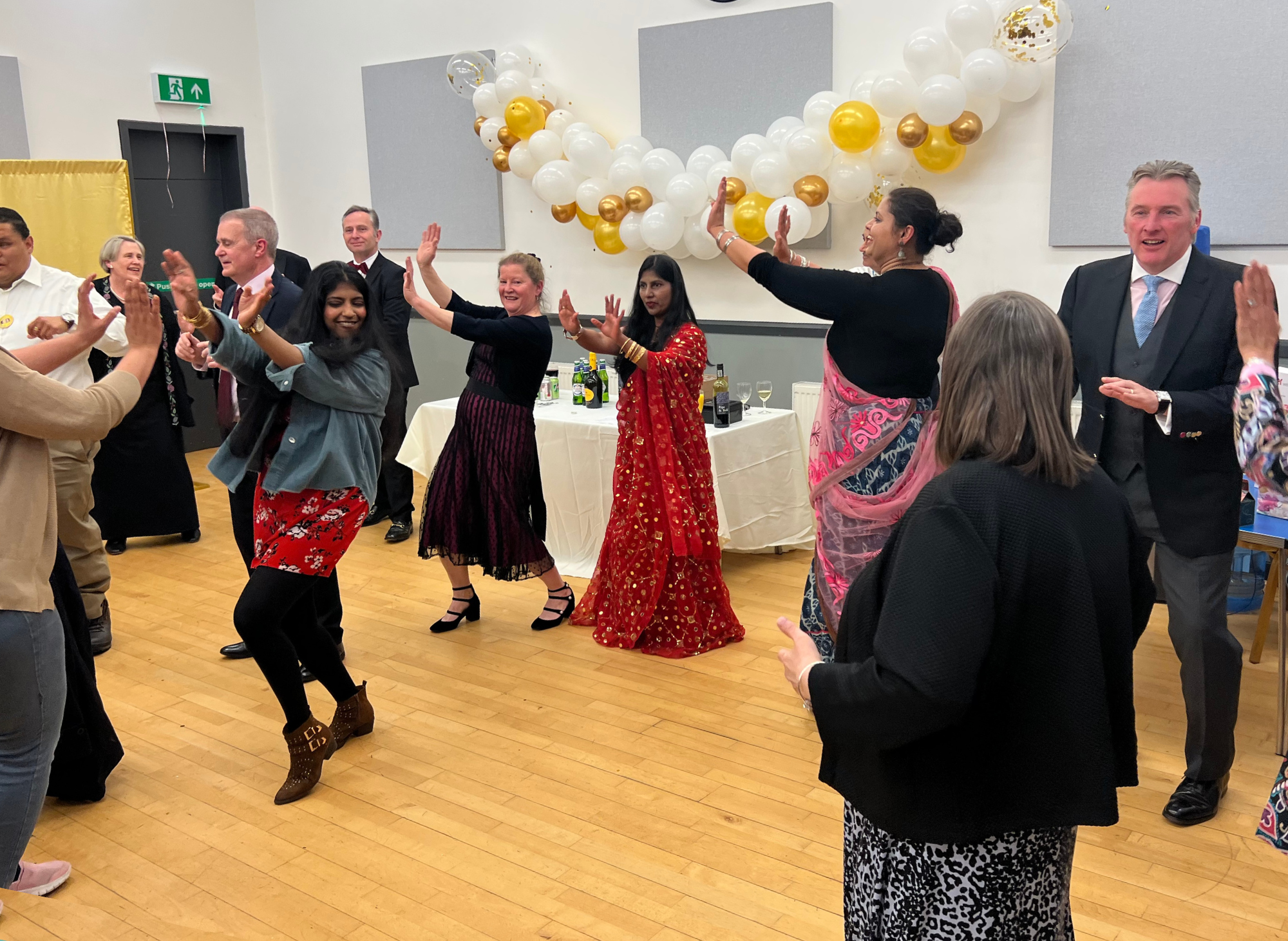 A hall full of people at Croydon Vision dancing together. Many are wearing special clothes and have their hands flung up to the left of their heads. The hall is decorated with white and gold balloons.