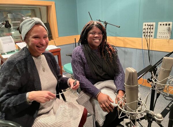 Two women sitting in a recording room, surrounded by recording equipment such as microphones