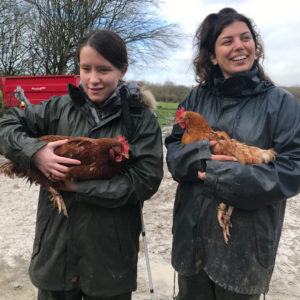 Two young women, Ellie and Naz, gently cradling chickens. They're wearing thick, black farm waterproofs and smiling.