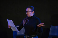 An actor from The Unseen holding a script and standing on stage. She has glasses, long, dark hair and is wearing a black turtle neck.