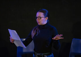 An actor from The Unseen holding a script and standing on stage. She has glasses, long, dark hair and is wearing a black turtle neck.